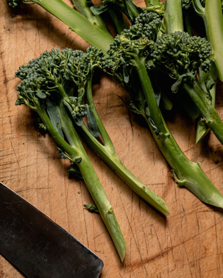 An overhead shot shows broccolini stalks on a wood cutting board. One has just been split in half lengthwise.