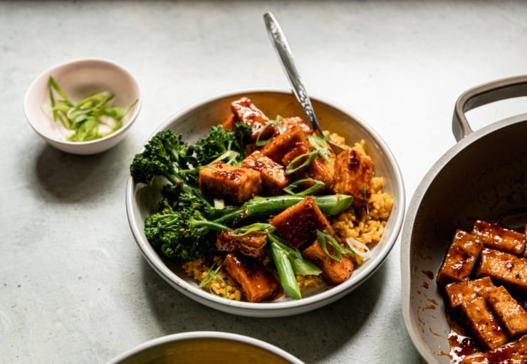 A 3/4 angle shot shows a spicy maple tofu bowl with golden garlic rice and spears of broccolini. A fork is sticking out of the bowl and it is garnished with sliced green onions.