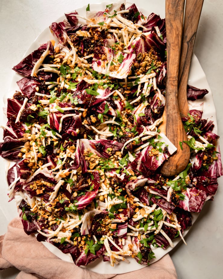 An overhead shot shows a platter with a celery root and radicchio salad topped with warm date dressing, chopped parsley, and chopped pistachios. Wood salad tongs are sticking out of the salad.