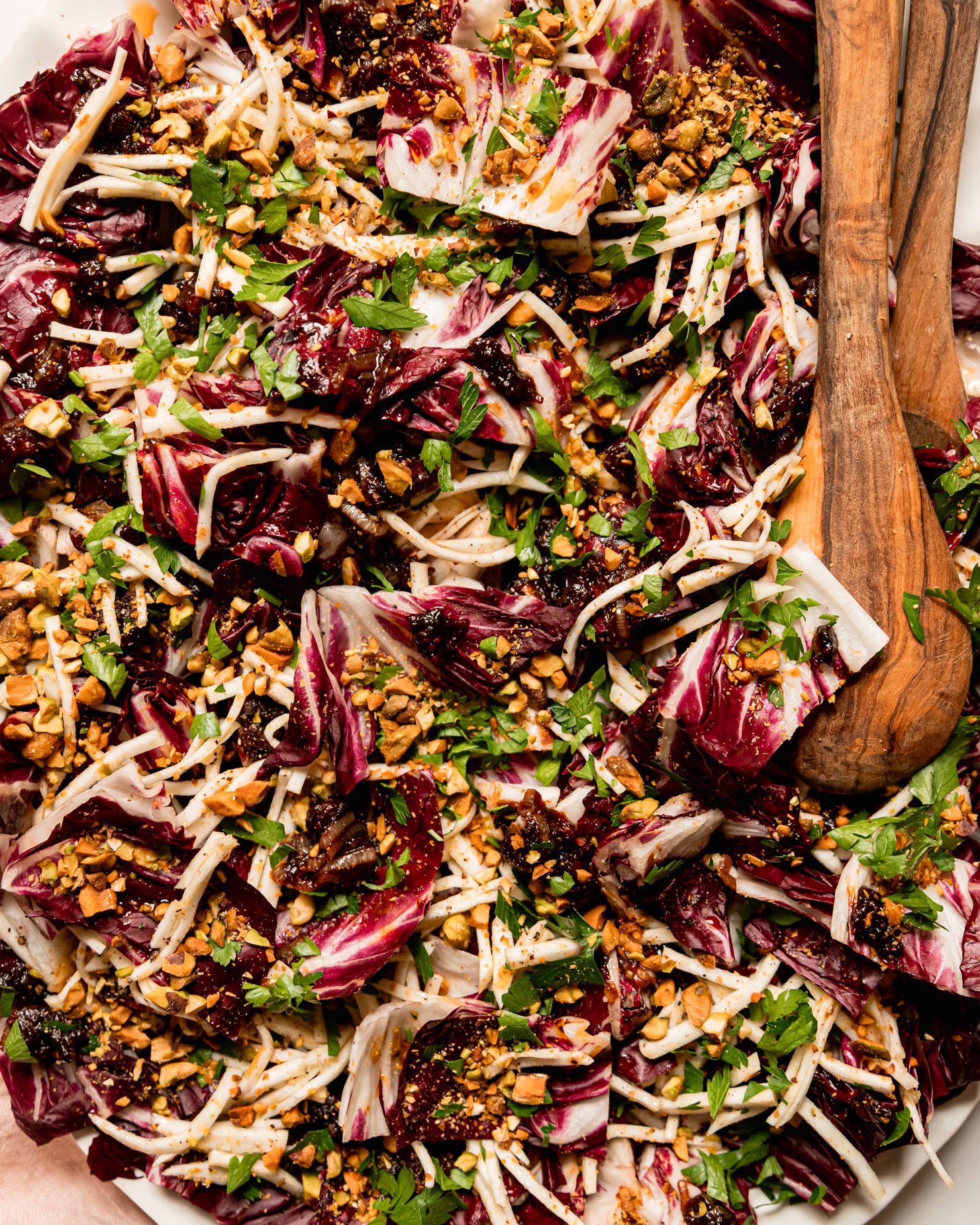An up close, overhead shot shows a platter with a celery root and radicchio salad topped with warm date dressing, chopped parsley, and chopped pistachios. Wood salad tongs are sticking out of the salad.