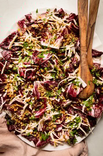 An overhead shot shows a platter with a celery root and radicchio salad topped with warm date dressing, chopped parsley, and chopped pistachios. Wood salad tongs are sticking out of the salad.