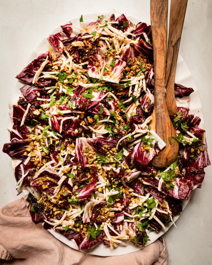 An overhead shot shows a platter with a celery root and radicchio salad topped with warm date dressing, chopped parsley, and chopped pistachios. Wood salad tongs are sticking out of the salad.