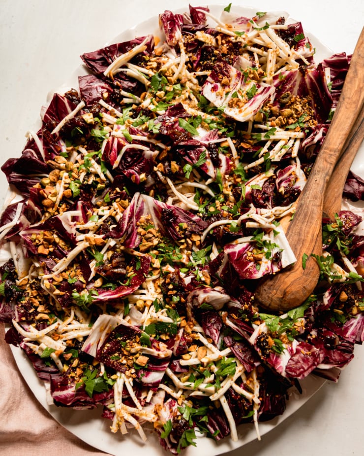 An overhead shot shows a platter with a celery root and radicchio salad topped with warm date dressing, chopped parsley, and chopped pistachios. Wood salad tongs are sticking out of the salad.