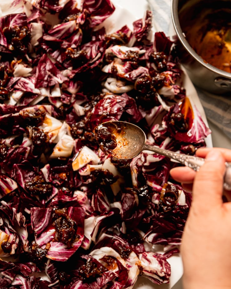 An overhead shot shows a hand spooning a warm date dressing on top of chopped radicchio leaves on a platter.
