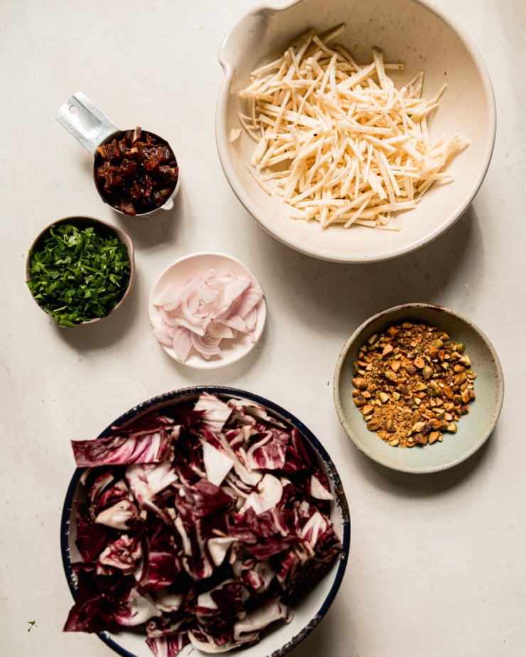 An overhead shot shows a bowl with julienned celery root, a bowl of chopped pistachios, a bowl with chopped radicchio, a small bowl of sliced shallots, a small bowl of chopped parsley, and a easuring cup with chopped dates.