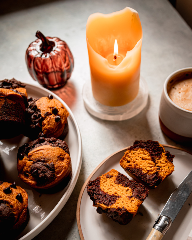 A 3/4 angle shot shows a plate of vegan pumpkin chocolate muffins, a glass pumpkin decoration, a lit candle, a cup of coffee, and a halved muffin on a plate, showing marbled chocolate and pumpkin batters.