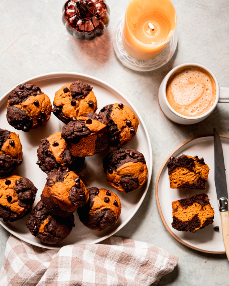 An overhead shot shows a plate of vegan pumpkin chocolate muffins, a glass pumpkin decoration, a lit candle, a cup of coffee, and a halved muffin on a plate, showing marbled chocolate and pumpkin batters.