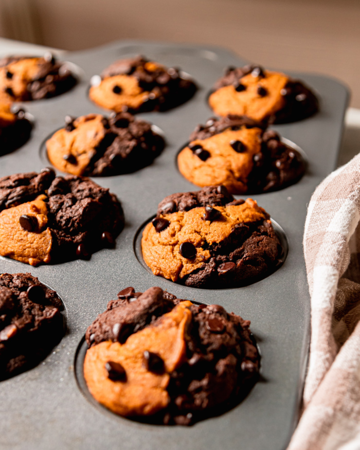 A 3/4 angle shot shows freshly baked marbled vegan pumpkin chocolate muffins in a muffin tin. The domes are puffed up nice and high and studded with chocolate chips.