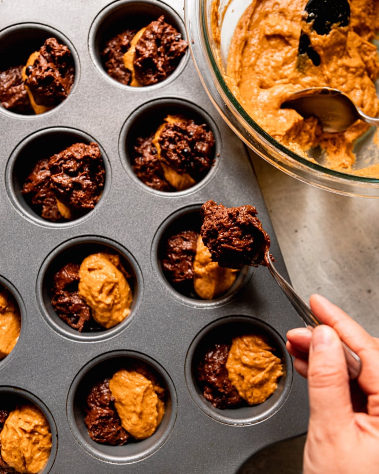 An overhead shot shows a hand portioning pumpkin and chocolate batters into a muffin tin.