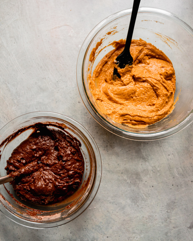 An overhead shot shows two bowls: one with a pumpkin batter and one with a chocolate batter. Both have spatulas sticking out of the bowls.