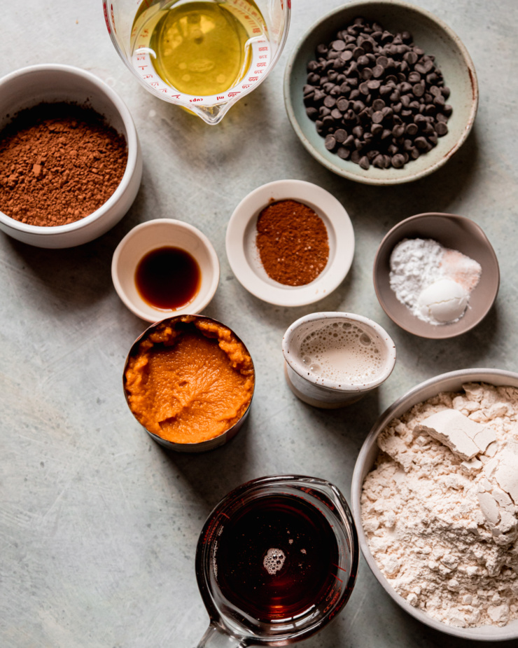 An overhead shot shows ingredients used for some vegan muffins: oil, cocoa powder, light spelt flour, pumpkin purée, maple syrup, spices, baking powder, baking soda, fine salt, vanilla, chocolate chips, and non-dairy milk.