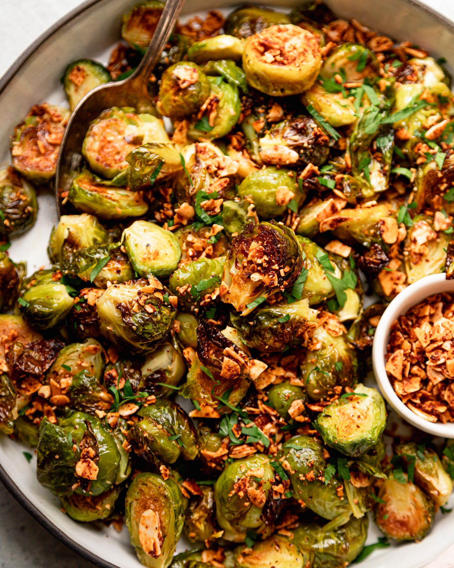 An up close, overhead shot shows a roasted Brussels sprouts dish covered in crushed up smoky maple almond bits and some chopped parsley.