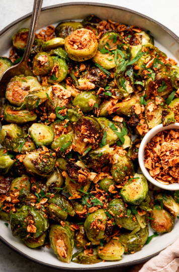 An up close, overhead shot shows a roasted Brussels sprouts dish covered in crushed up smoky maple almond bits and some chopped parsley.