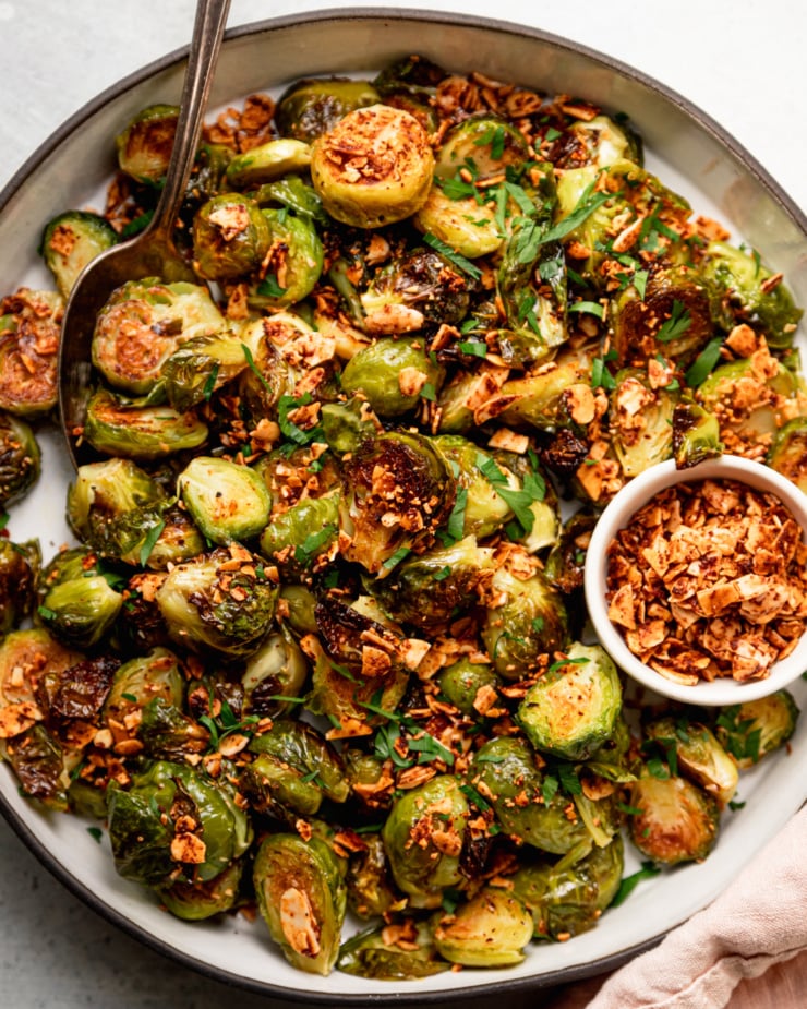 An up close, overhead shot shows a roasted Brussels sprouts dish covered in crushed up smoky maple almond bits and some chopped parsley.