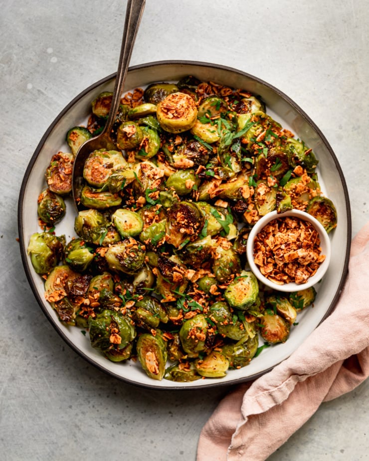 An overhead shot shows a roasted Brussels sprouts dish covered in crushed up smoky maple almond bits and some chopped parsley.