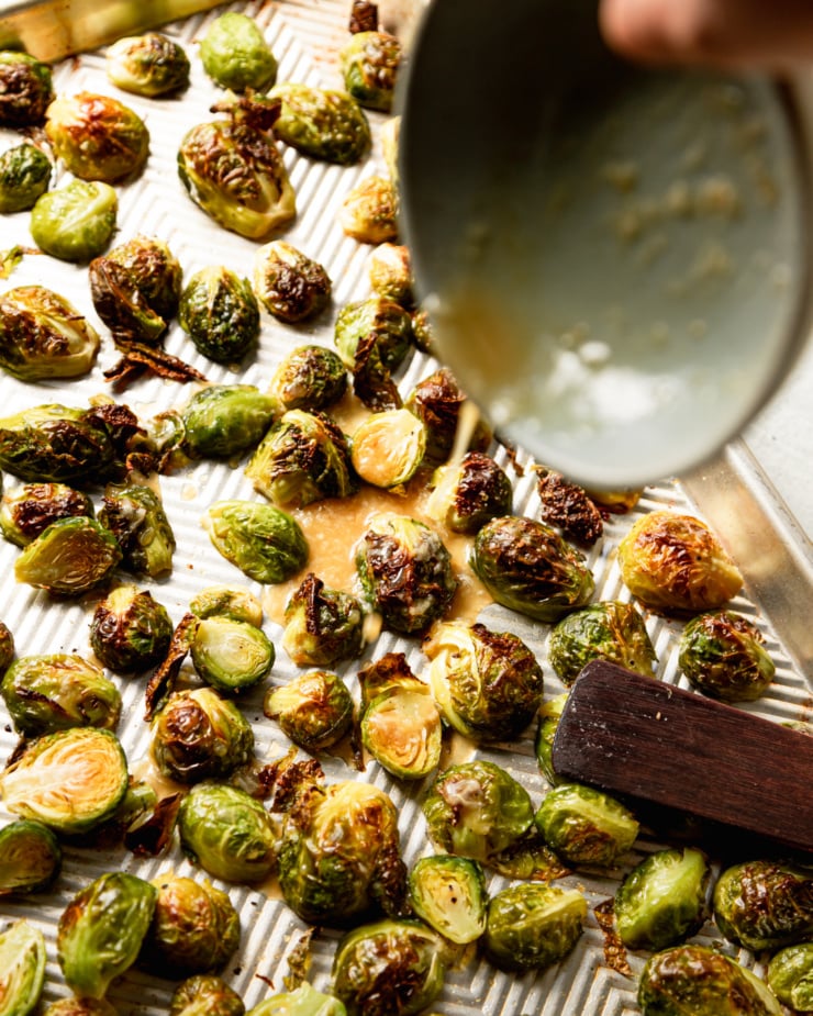 An overhead shot shows a hand using a bowl to pour a lemon juice/maple/garlic/Dijon mustard mixture over roasted brussels sprouts.