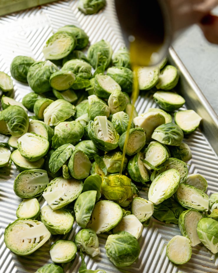 An overhead shot shows olive oil being poured over halved Brussels sprouts.