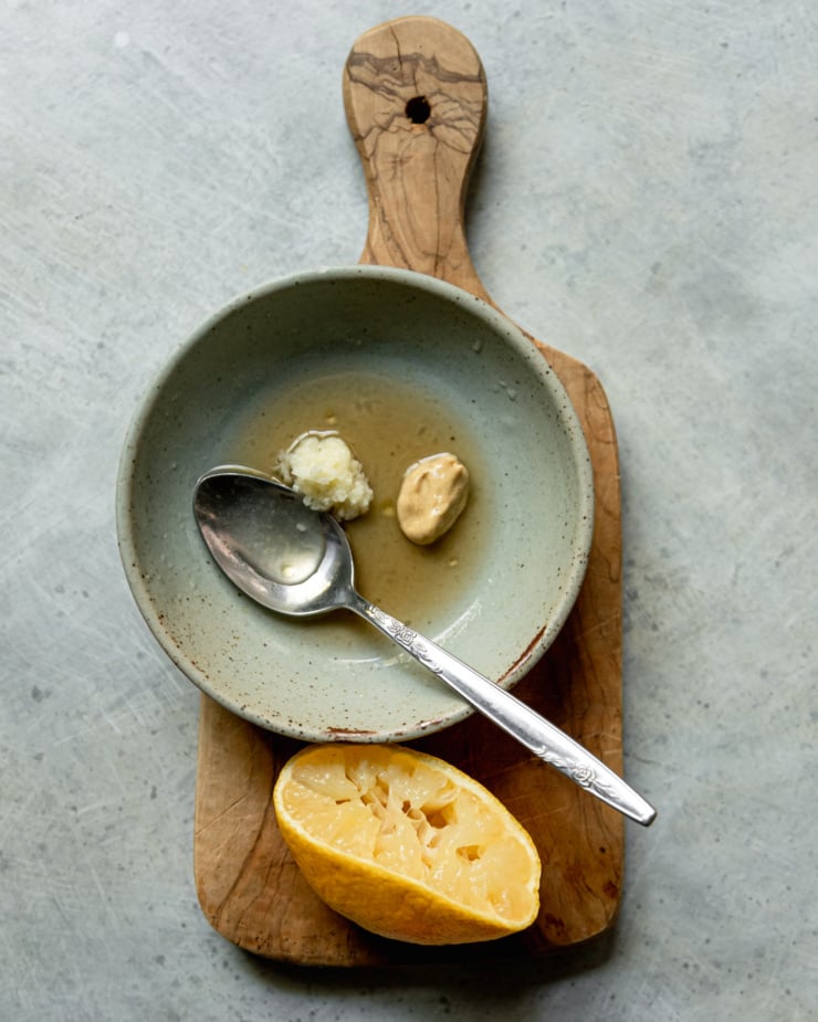 An overhead shot shows a small bowl and squeezed lemon half on a small wooden cutting board. In the bowl is lemon juice, maple syrup, finely grated garlic, and Dijon mustard.