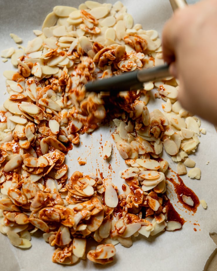 An overhead shot shows a hand using a spatula to stir a spice/maple/oil mixture into some sliced almonds.