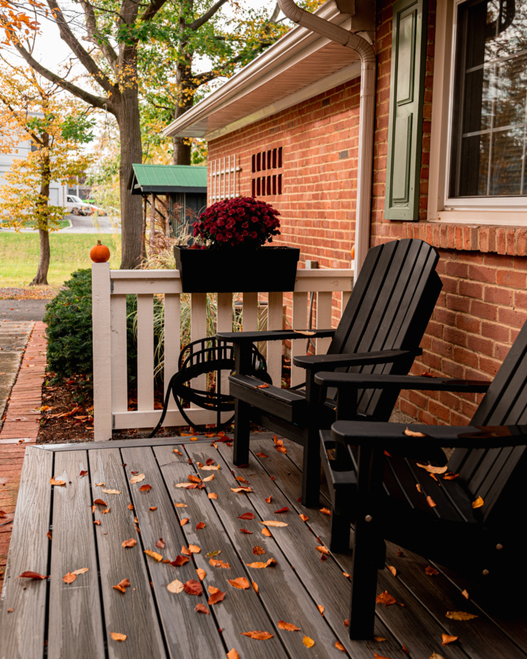 A head-on shot shows a porch with two dark deck chairs and autumn leaves all on the ground. A white fence with a chrysanthemum planter on top and a brick house is seen in the background.