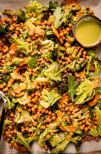 An up close, overhead shot shows a baking sheet salad with roasted delicata squash and broccoli, chickpeas, bibb lettuce, baby arugula, walnuts, and creamy shallot herb dressing. A small bowl of dressing is to the side and a set of salad tongs is sticking out of the salad.