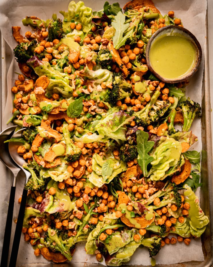 An up close, overhead shot shows a baking sheet salad with roasted delicata squash and broccoli, chickpeas, bibb lettuce, baby arugula, walnuts, and creamy shallot herb dressing. A small bowl of dressing is to the side and a set of salad tongs is sticking out of the salad.