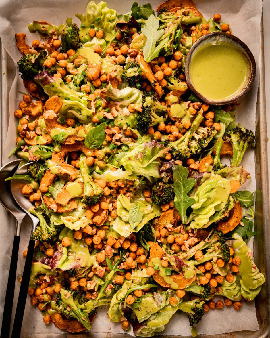 An up close, overhead shot shows a baking sheet salad with roasted delicata squash and broccoli, chickpeas, bibb lettuce, baby arugula, walnuts, and creamy shallot herb dressing. A small bowl of dressing is to the side and a set of salad tongs is sticking out of the salad.