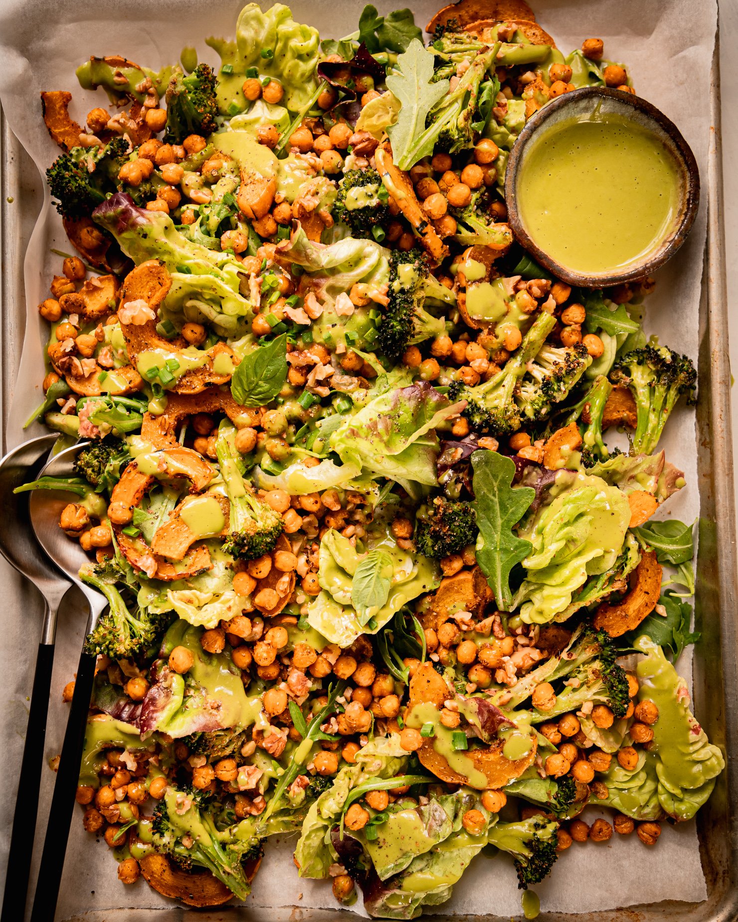 An up close, overhead shot shows a baking sheet salad with roasted delicata squash and broccoli, chickpeas, bibb lettuce, baby arugula, walnuts, and creamy shallot herb dressing. A small bowl of dressing is to the side and a set of salad tongs is sticking out of the salad.