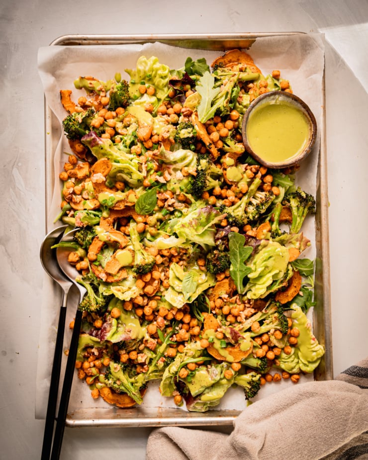 An overhead shot shows a baking sheet salad with roasted delicata squash and broccoli, chickpeas, bibb lettuce, baby arugula, walnuts, and creamy shallot herb dressing. A small bowl of dressing is to the side and a set of salad tongs is sticking out of the salad.