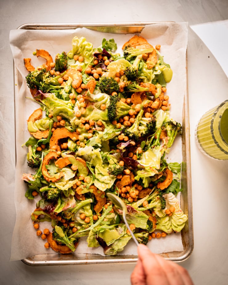 An overhead shot shows a hand using a spoon to drizzle creamy shallot herb dressing onto a baking sheet salad.