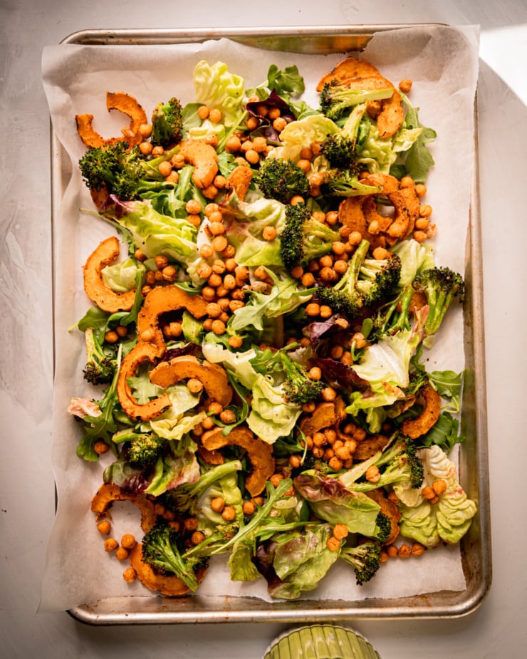 An overhead shot shows a baking sheet salad with bibb lettuce, arugula, roasted delicata squash, roasted broccoli, and chickpeas.