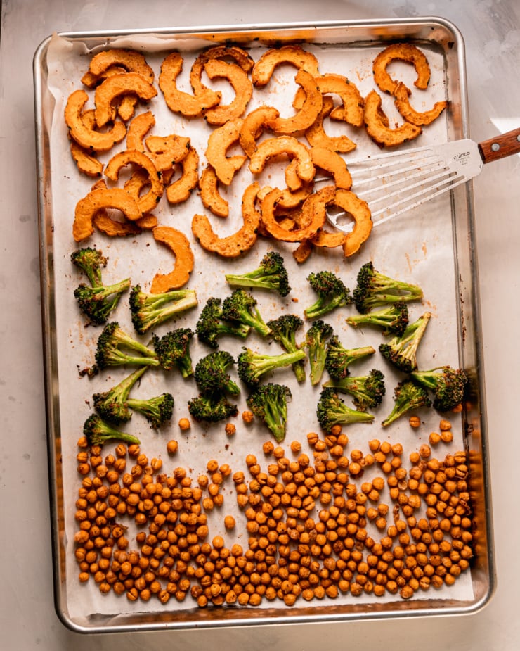 An overhead shot shows a parchment lined baking sheet with roasted delicata squash slices, roasted broccoli, and chickpeas.