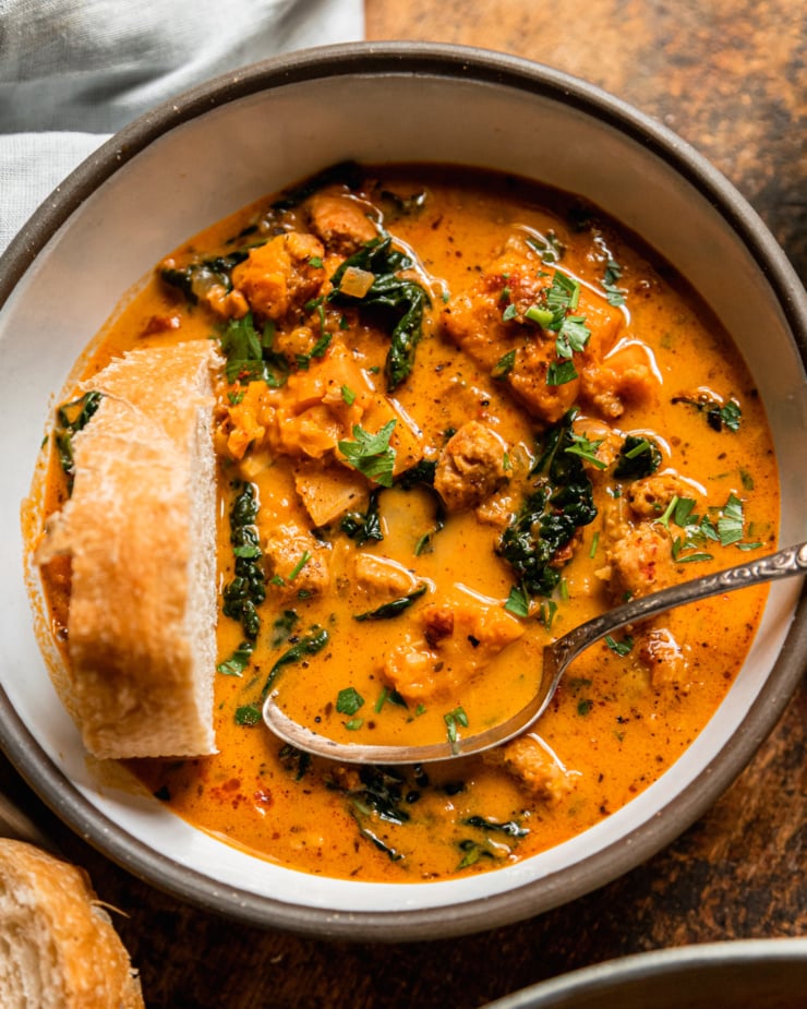 An up close, overhead shot shows a bowl of vegan butternut squash zuppa Toscana. The soup is creamy and pale orange in hue with chopped parsley, bits of cooked vegan veggie sausage, and kale. An antique spoon and a piece of crusty bread are sticking out of the bowl.