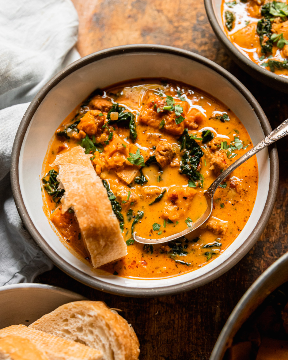 An overhead shot shows a bowl of vegan butternut squash zuppa Toscana. The soup is creamy and pale orange in hue with chopped parsley, bits of cooked vegan veggie sausage, and kale. An antique spoon and a piece of crusty bread are sticking out of the bowl.