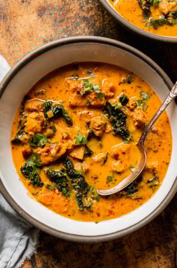 An overhead shot shows a bowl of vegan butternut squash zuppa Toscana. The soup is creamy and pale orange in hue with chopped parsley, bits of cooked vegan veggie sausage, and kale. An antique spoon is sticking out of the bowl.