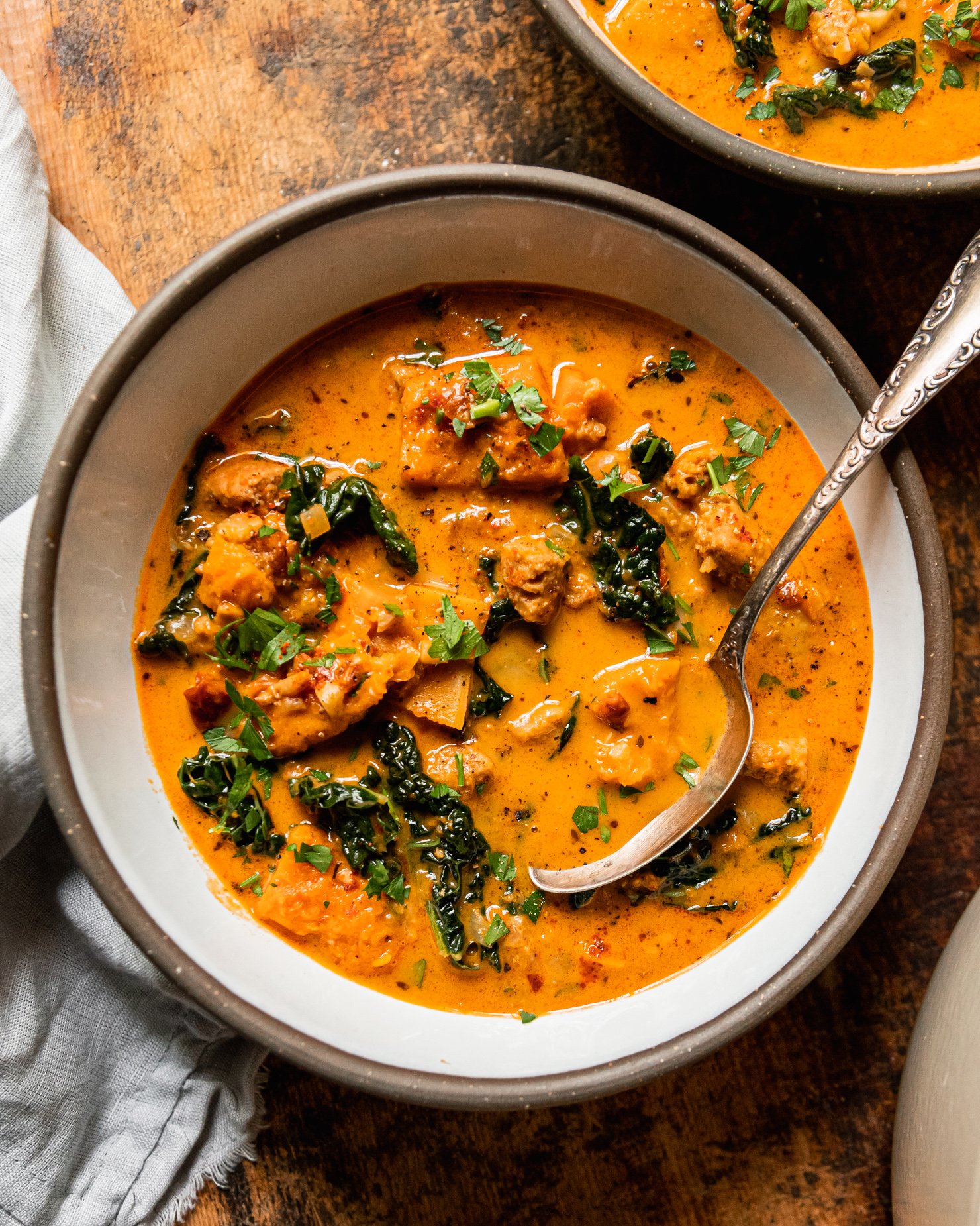 An overhead shot shows a bowl of vegan butternut squash zuppa Toscana. The soup is creamy and pale orange in hue with chopped parsley, bits of cooked vegan veggie sausage, and kale. An antique spoon is sticking out of the bowl.