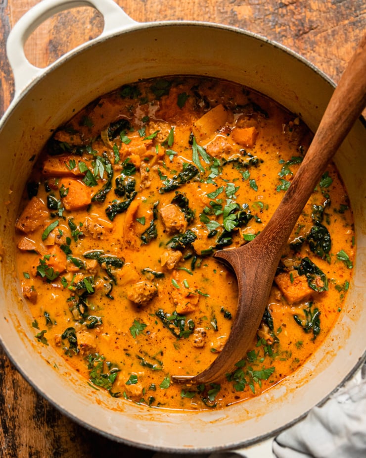An overhead shot shows a pot of vegan butternut squash zuppa Toscana. The soup is creamy and pale orange in hue with chopped parsley, bits of cooked vegan veggie sausage, and kale. A wooden spoon is sticking out of the pot.