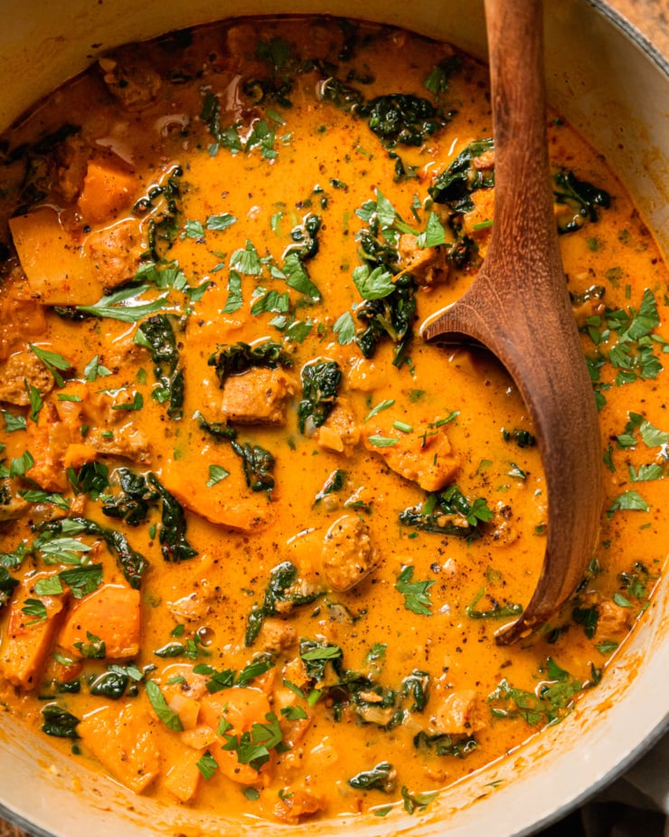 An up close, overhead shot shows a pot of vegan butternut squash zuppa Toscana. The soup is creamy and pale orange in hue with chopped parsley, bits of cooked vegan veggie sausage, and kale. A wooden spoon is sticking out of the pot.