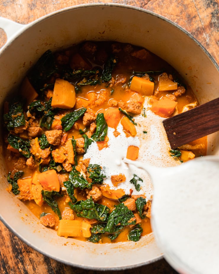 An overhead shot shows bright white cashew cream being poured into a pot of vegan butternut squash zuppa Toscana