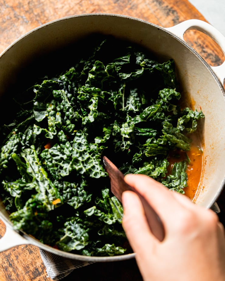An overhead shot shows a hand using a wooden utensil to stir chopped kale into a pot of soup.