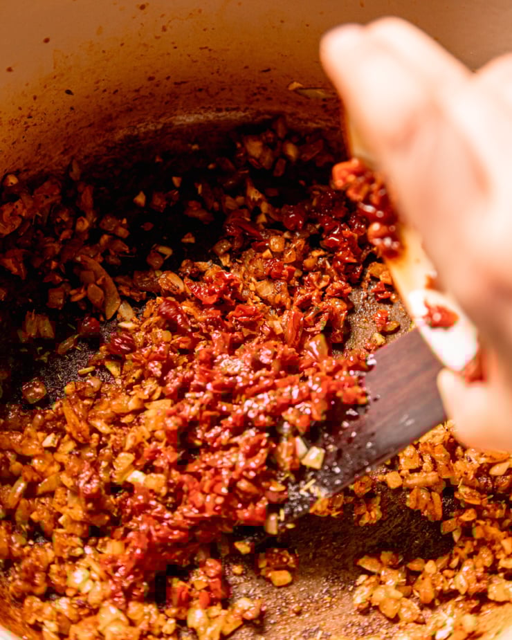 An overhead shot shows a hand emptying a bowl if minced sun dried tomatoes into a pot with sautรฉed onions and spices.