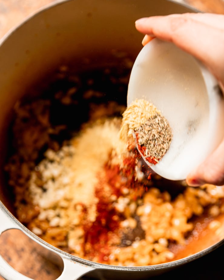 An overhead shot shows a hand emptying a bowl of spices into a pot with sautรฉed onions.