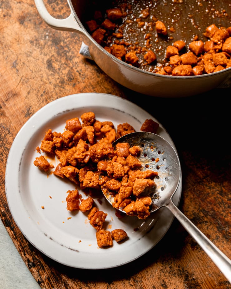 An overhead shot shows a slotted spoon transferring cooked vegan veggie sausage pieces to a plate from a pot.