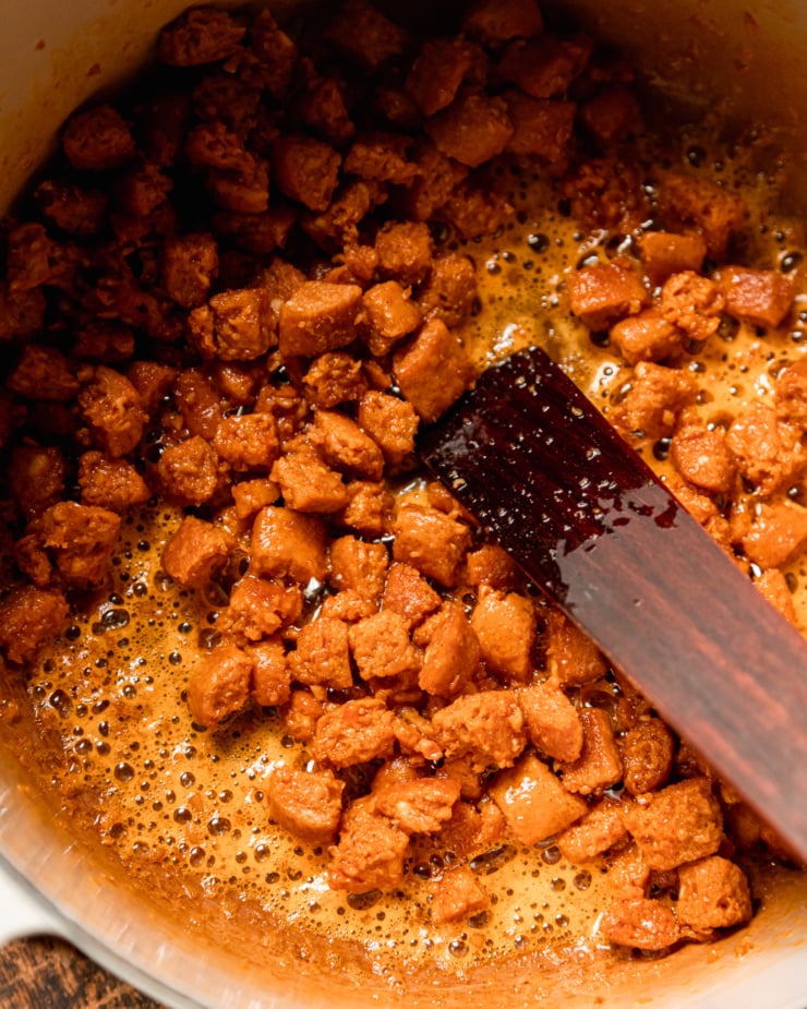 An overhead shot shows vegan veggie sausage pieces sizzling in a pot with a wooden spatula.