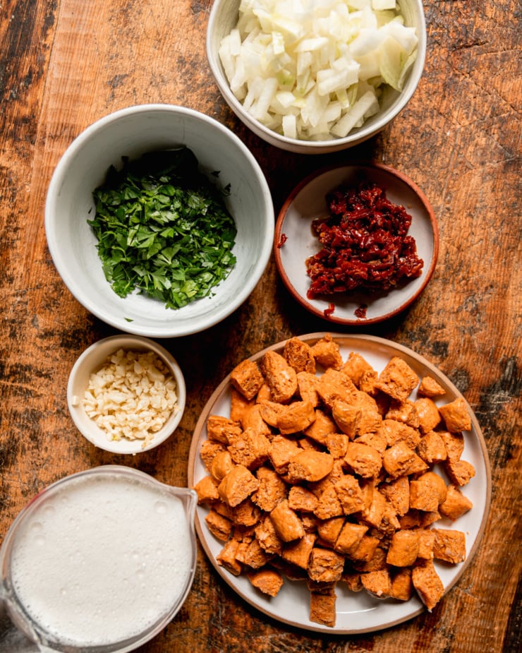 An overhead shot shows prepped ingredients for a vegan soup: chopped veggie sausage, cashew cream, minced garlic, chopped parsley, chopped onions, and minced sun dried tomatoes; all on a rough wooden board.