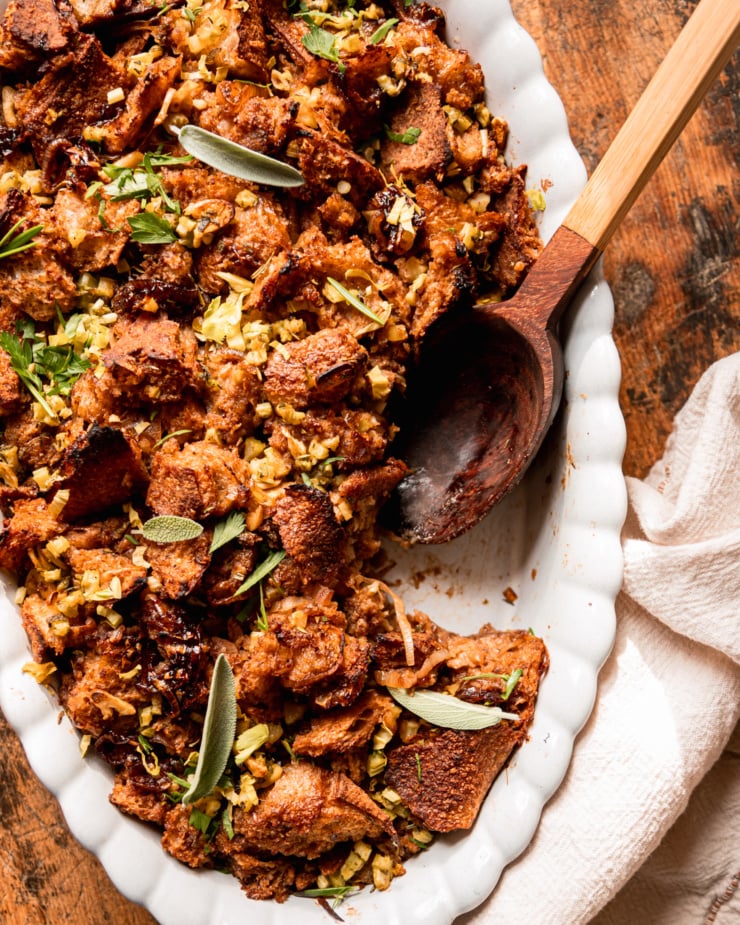 An up close overhead shot shows a dish of baked vegan stuffing with rustically torn sourdough bread, balsamic shallots, celery, and herbs. A wooden serving spoon is sticking out of the stuffing dish.