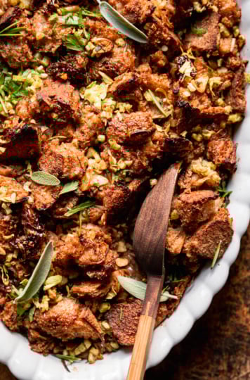 An up close overhead shot shows a dish of baked vegan stuffing with rustically torn sourdough bread, balsamic shallots, celery, and herbs. A wooden serving spoon is sticking out of the stuffing dish.