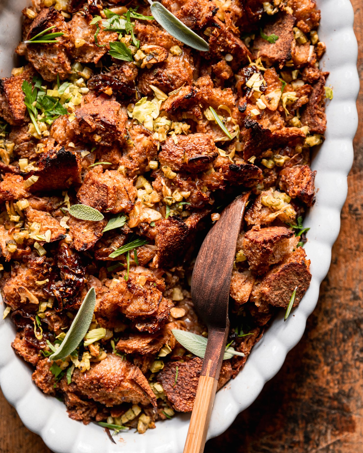 An up close overhead shot shows a dish of baked vegan stuffing with rustically torn sourdough bread, balsamic shallots, celery, and herbs. A wooden serving spoon is sticking out of the stuffing dish.