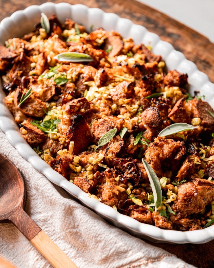 A 3/4 angle shot shows a baking dish with vegan stuffing on a rough wooden board surface. Herbs garnish the top.