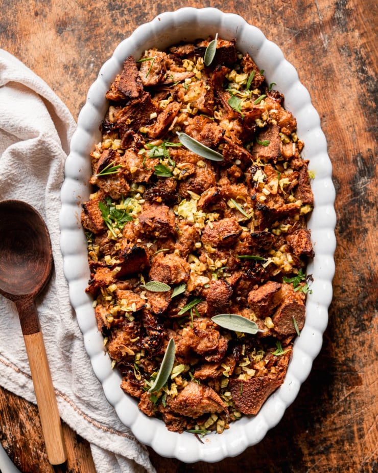 An overhead shot shows a dish of baked vegan stuffing with rustically torn sourdough bread, balsamic shallots, celery, and herbs. A wooden serving spoon is seen to the side.